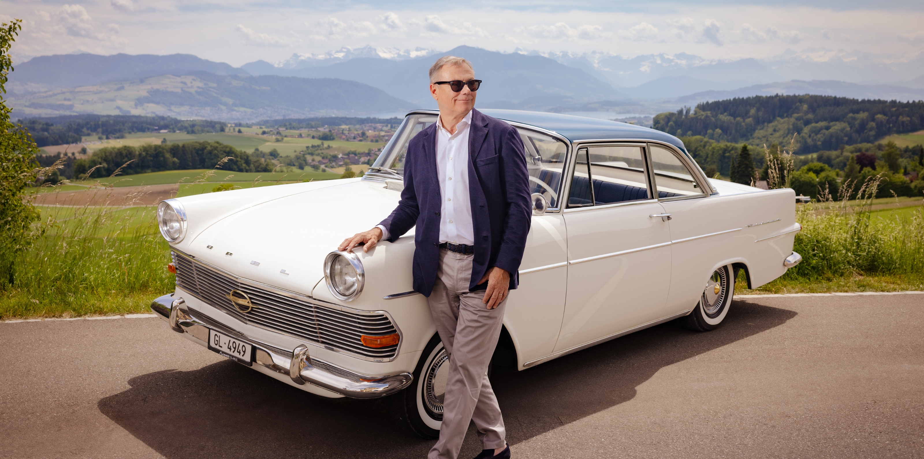 Klassik und Landschaft Mann lehnt an einem klassischen weissen Auto mit malerischem Alpenhintergrund.